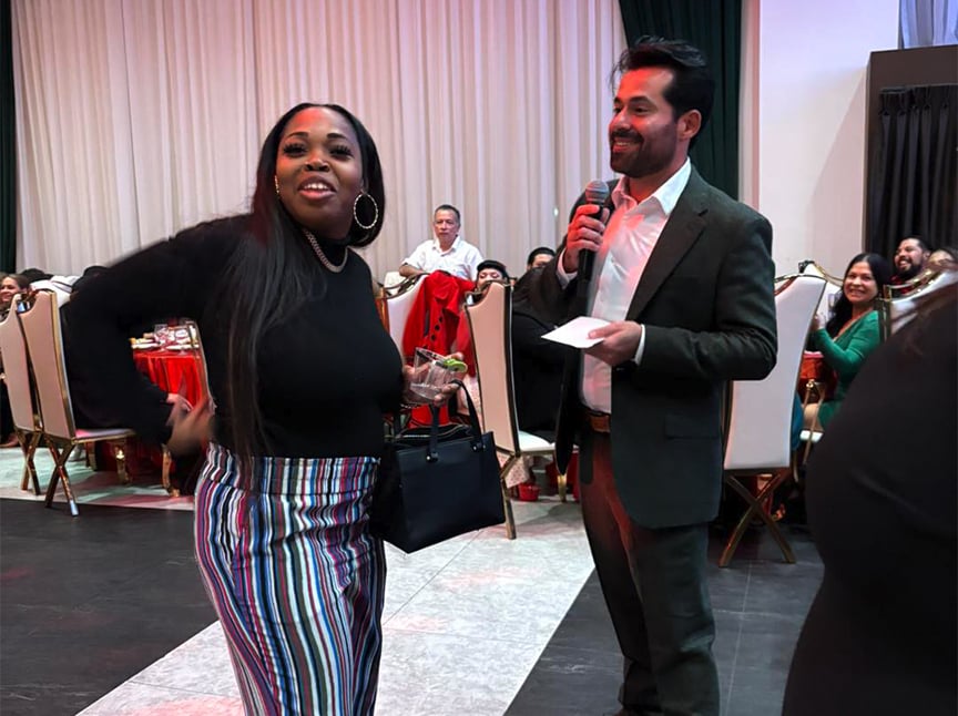 A woman in a black top and striped pants smiles while holding a drink and purse, as a man in a suit with a microphone and paper stands beside her. People are seated and watching in a decorated event hall.