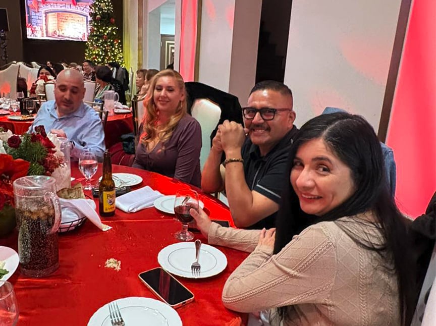 Four people sit at a red-decorated table, smiling and enjoying drinks at a festive indoor gathering with a Christmas tree and lights visible in the background. White plates and glasses are on the table.