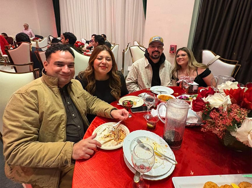 Four people sit around a red tablecloth at a restaurant, smiling at the camera. Plates with leftover food, glasses, and a floral centerpiece are on the table. Other guests are visible in the background.