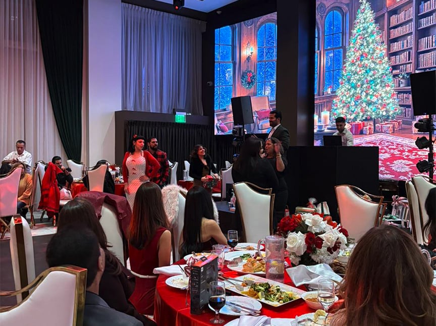 A festive indoor event with people seated at tables, enjoying food and drinks. A large digital backdrop shows a decorated Christmas tree and a fireplace. Some guests stand and interact near the front of the room.