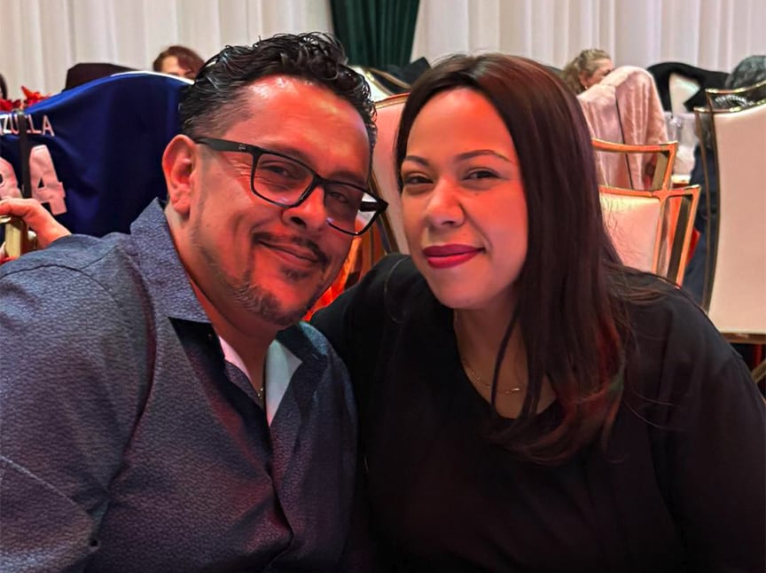 A man wearing glasses and a blue shirt sits closely beside a woman with long brown hair and a black top. They are both smiling at the camera in a warmly lit indoor setting.