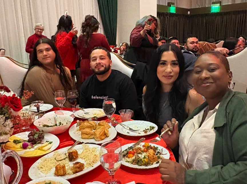 Four people sit at a red-covered table, smiling and enjoying a meal together at a festive indoor gathering. Plates of food and drinks are on the table, with more people and decorations in the background.