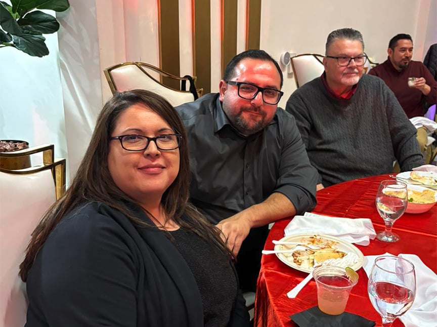 Three people sit at a table covered with a red tablecloth, enjoying food and drinks. They are smiling and wearing glasses. The setting appears to be a formal or festive gathering.