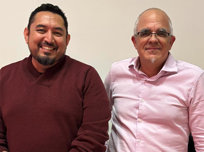 Two men are standing side by side against a plain light-colored wall. The man on the left is smiling, wearing a maroon sweater, and the man on the right is wearing glasses and a light pink shirt, also smiling.