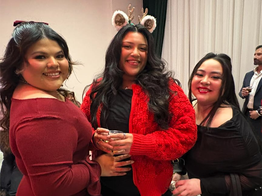 Three women smile and pose together at a social event. The woman in the center wears a red sweater and reindeer antler headband. All appear cheerful in a festive indoor setting.