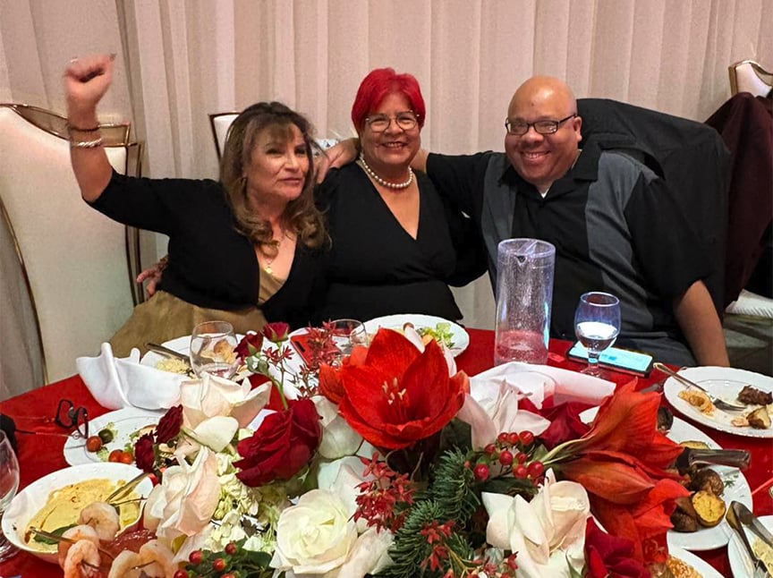 Three adults sit together at a table decorated with red and white flowers, smiling and posing for a photo during what appears to be a festive meal or celebration. Plates of food and drinks are in front of them.