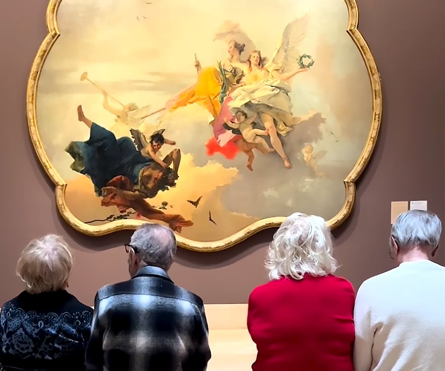 Four elderly people sit on a bench facing a large, ornate-framed painting of four winged, mythological figures floating in the sky, displayed in a museum setting.