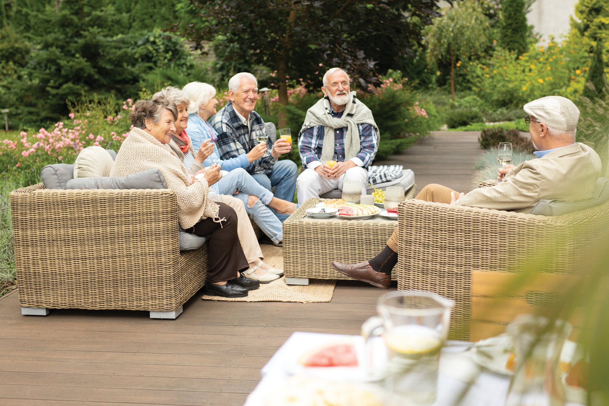A group of six older adults sit on wicker patio furniture outdoors, enjoying drinks and conversation. They are surrounded by lush greenery and flowers, creating a relaxed, cheerful atmosphere.