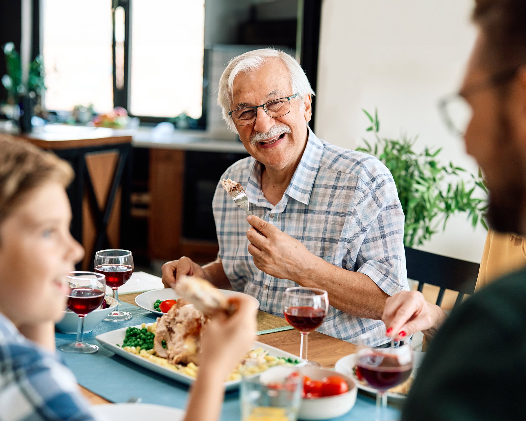 An elderly man with white hair and glasses smiles while eating at a dinner table with family. Plates of food and glasses of red drink are on the table, and the atmosphere is warm and cheerful.