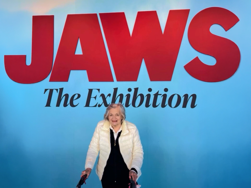 An elderly woman in a white jacket smiles while standing in front of a blue wall with large red text that reads JAWS and smaller black text below that says The Exhibition.