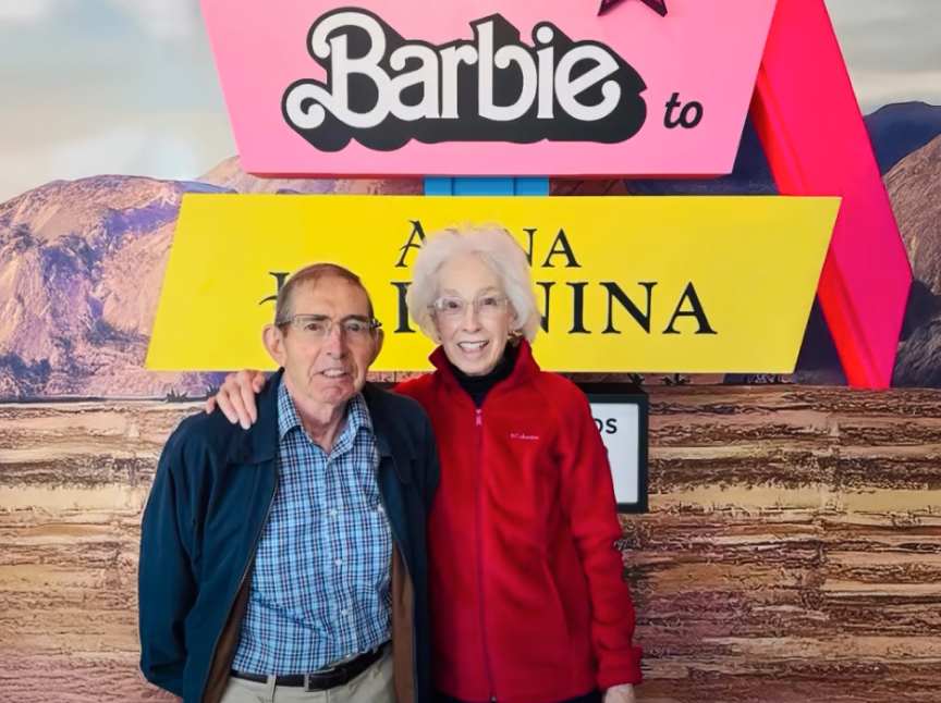 An older man and woman smile while standing arm-in-arm in front of a colorful “Barbie” sign with a desert mountain backdrop. The man wears glasses, a blue jacket, and plaid shirt; the woman wears glasses and a red jacket.
