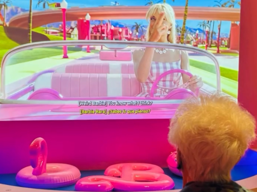 A person with white hair sits facing a large screen showing a blonde woman dressed in pink driving a pink convertible in a colorful, cartoon-like world. The scene has subtitles in English and Spanish.