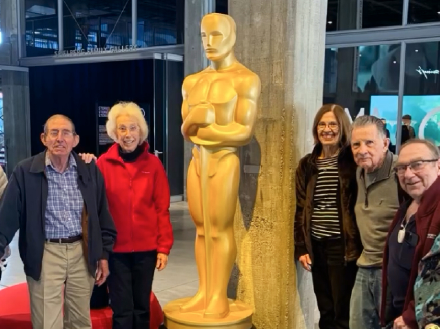 A group of five smiling older adults stands beside a large gold Oscar statue in an indoor setting with glass walls and modern decor.