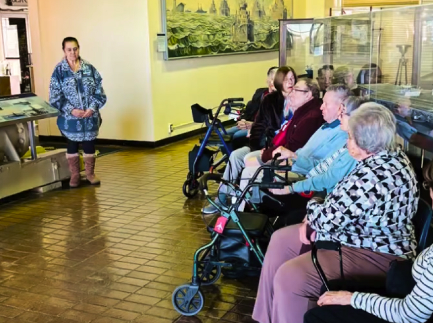 A group of elderly people sits in chairs, some with walkers, listening to a woman standing and speaking in a brightly lit museum or gallery room.