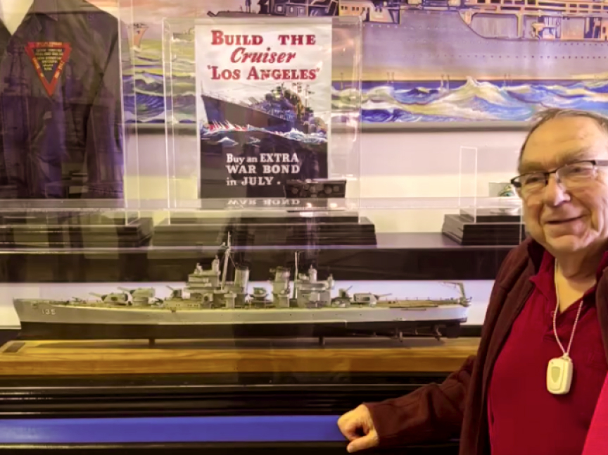 A smiling man stands beside a display case containing a model warship. Behind the case is a vintage Build the Cruiser Los Angeles war bonds poster and a ship painting. The man wears glasses and a red shirt.