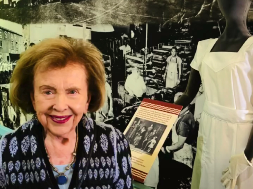 An elderly woman with brown hair smiles in front of an exhibit featuring a mannequin in a white dress and a historical black-and-white photo backdrop. An informational display stands beside the mannequin.