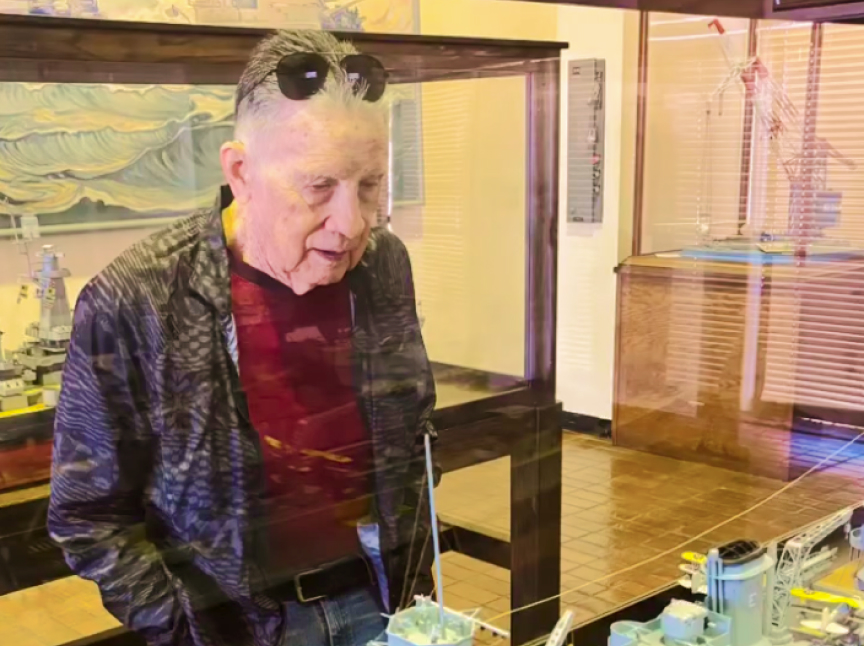 An older man wearing sunglasses on his head and a red shirt looks at a detailed ship model inside a glass display case in a museum or exhibit room.