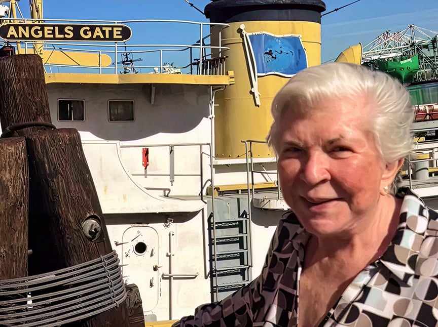 An older woman with short white hair smiles in front of a yellow and white ship named Angels Gate, docked at a harbor on a sunny day.