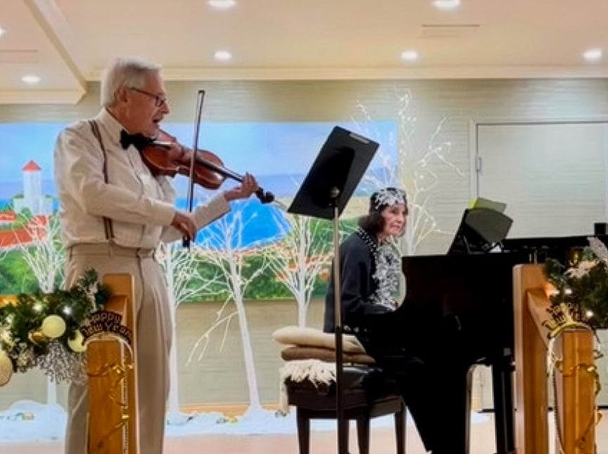 An elderly man in a white shirt and bow tie plays violin beside a smiling elderly woman at a piano. The room is decorated with festive “Happy New Year” signs and garlands, and a painted mural is on the back wall.
