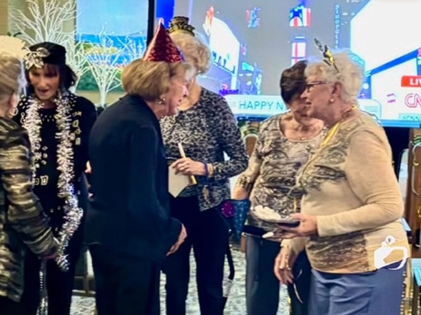 A group of elderly women in festive hats and accessories gather indoors, smiling and talking, celebrating at a New Year’s event with party decorations and a TV in the background.
