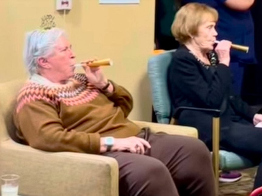 Two older adults sit side by side indoors, each blowing on a gold party horn. One wears a festive headband and a patterned sweater, suggesting a celebratory occasion. Both appear relaxed and focused on the activity.
