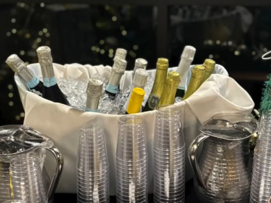 A white cloth-lined bucket filled with ice chills bottles of champagne and sparkling wine. Plastic cups are stacked in front, alongside pitchers of water. Warm lights are blurred in the background.