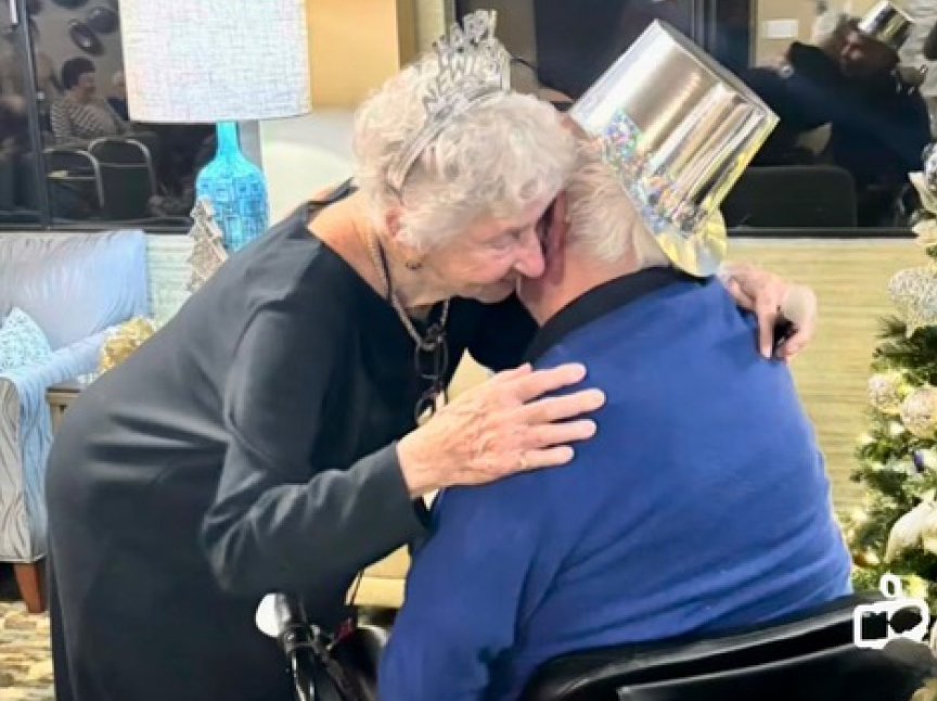 An elderly woman wearing a tiara hugs an elderly man in a festive hat. They are celebrating near a decorated Christmas tree, sharing a warm, emotional embrace.