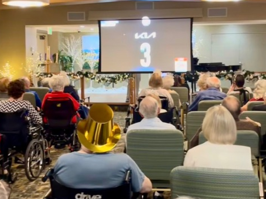 A group of seniors, some in wheelchairs, sit in a decorated room watching a large screen. One person wears a shiny gold hat. The room has festive lights and holiday decor, creating a cheerful atmosphere.