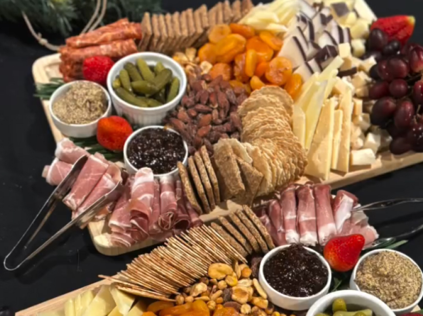 A charcuterie board with assorted cheeses, crackers, prosciutto, dried apricots, grapes, strawberries, almonds, pickles, nuts, breadsticks, meats, and small bowls of mustard and jam.