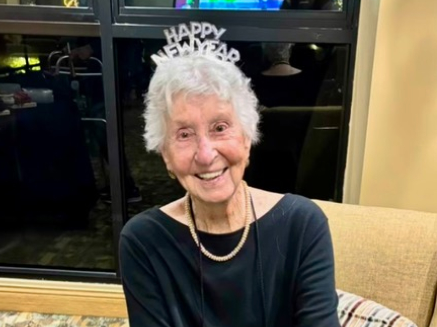 Elderly woman with short white hair smiles while wearing a Happy New Year headband and a black top with a pearl necklace, sitting on a couch in front of a window.