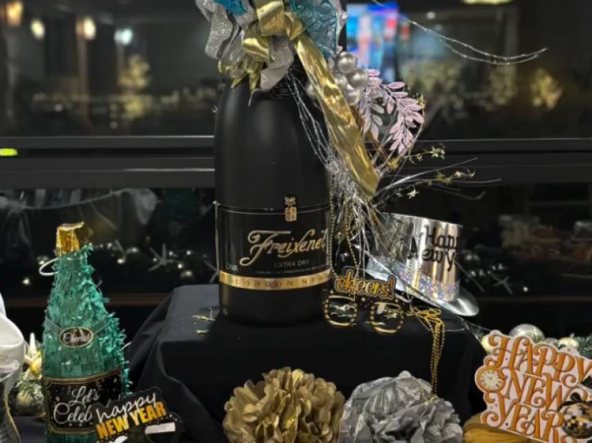 A decorated table displays a large Freixenet champagne bottle surrounded by gold and silver New Year’s decorations, party glasses labeled “cheers,” and signs reading “Happy New Year.”.