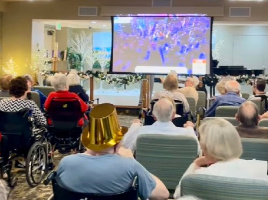 A group of seniors, some in wheelchairs, sit facing a large screen showing a colorful event. One person wears a shiny gold party hat. The room is decorated with festive lights and garlands.