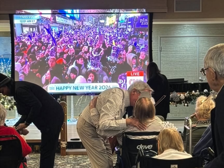 A group of elderly people celebrate New Years Eve in a room, watching a TV broadcast of a large crowd in Times Square. A man hugs a woman in a wheelchair, while others talk and watch the screen. “Happy New Year 2024” is displayed on TV.