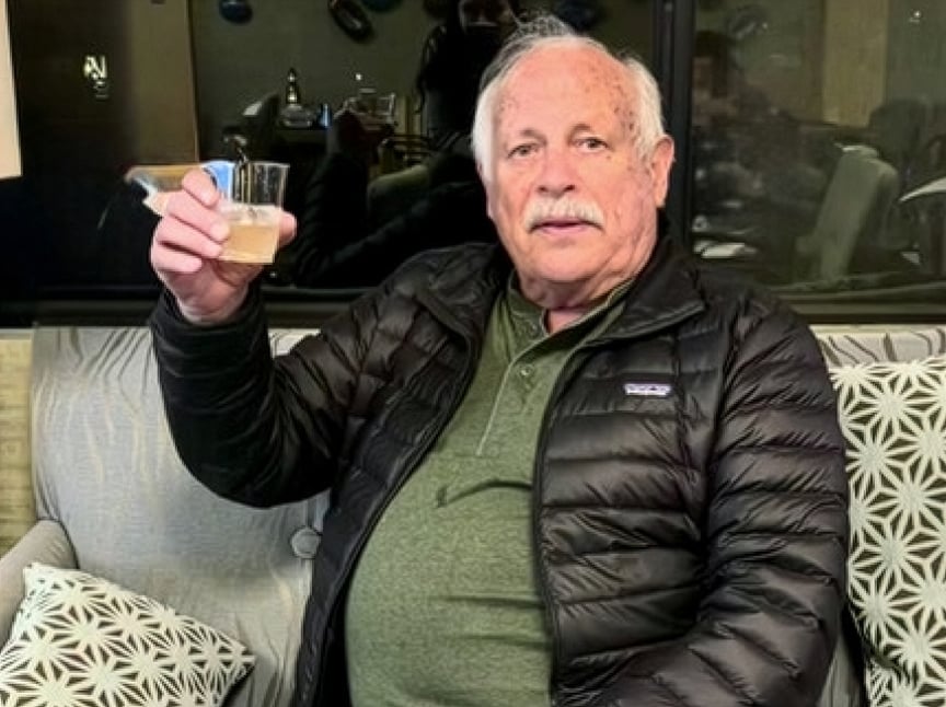 An older man with gray hair and a mustache sits on a couch, wearing a green shirt and black puffer jacket, holding up a drink and looking at the camera. Patterned pillows are beside him.