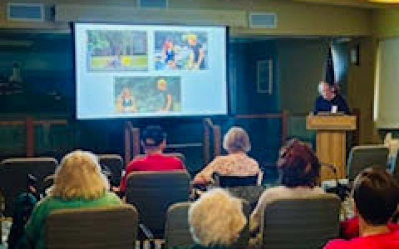 A speaker stands at a podium presenting to an audience seated in rows, with a large screen displaying four outdoor photos at the front of the room.