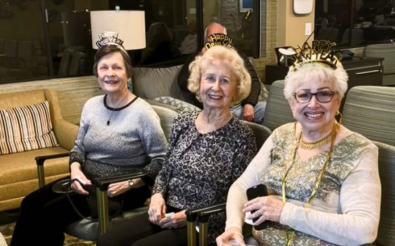 Three older women wearing Happy New Year headbands and festive clothes sit together smiling in a living room, celebrating New Year’s Eve. A man is seated behind them, and the atmosphere is cheerful and cozy.