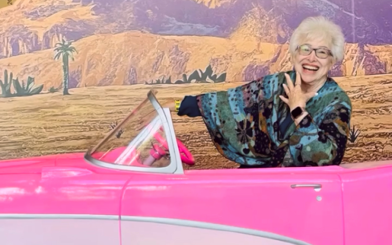 An older woman with white hair and glasses smiles joyfully while posing in a bright pink convertible car, set against a colorful desert mural background.