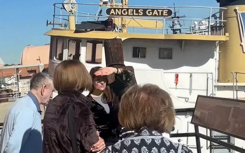 A group of four people stands on a dock near a boat named “Angels Gate.” One woman shades her eyes from the sun, while others face her. The boats deck and railing are visible in the background.