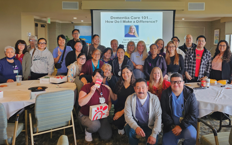 A large group of people smile and pose together indoors around tables with food. A projected slide behind them reads “Dementia Care 101… How Do I Make a Difference?” with a photo of a speaker.