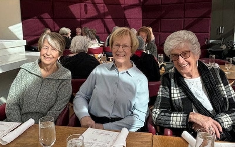 Three older women sit together at a restaurant table, smiling at the camera. Sunlight streams in, illuminating the group. There are menus, glasses of water, and napkins on the wooden table in front of them.