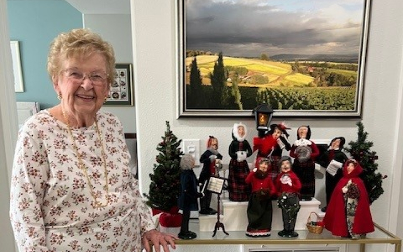 An elderly woman smiles beside a table decorated with festive Christmas carol figurines and small trees, with a landscape painting hanging on the wall behind her.
