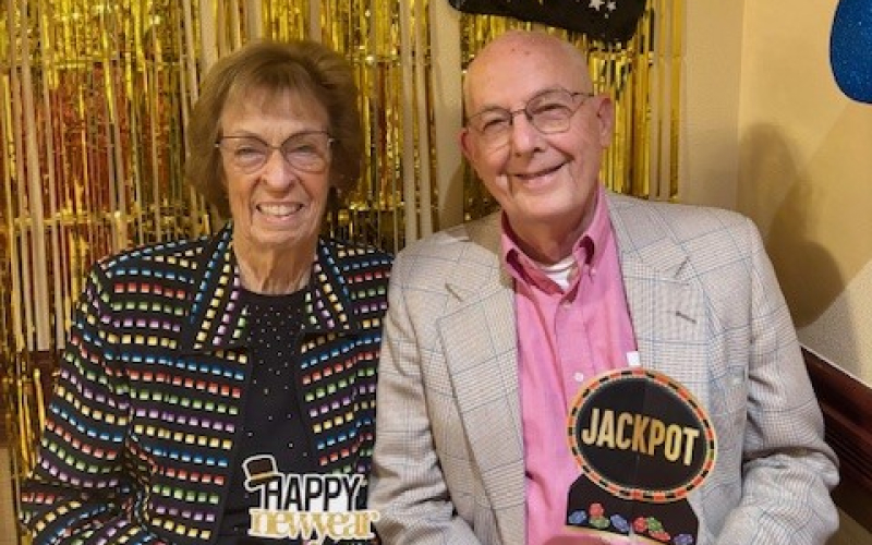 Two smiling older adults sit side by side at a party, holding signs that say Happy New Year and Jackpot. They wear festive outfits, with a gold tinsel backdrop behind them.