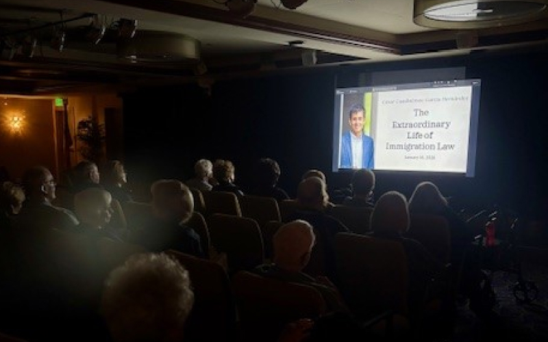 An audience sits in a dimly lit room watching a projected presentation titled The Extraordinary Life of Immigration Law with a speaker’s photo and the date, January 16, 2020, displayed on the screen.