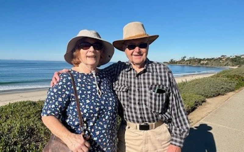 An older couple wearing hats and sunglasses stand arm in arm, smiling on a sunny coastal walkway with the ocean and blue sky in the background.