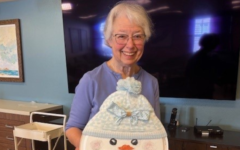 An older woman with short gray hair and glasses smiles while holding a snowman decoration wearing a blue knit hat and bow. She stands indoors in front of a large TV screen.
