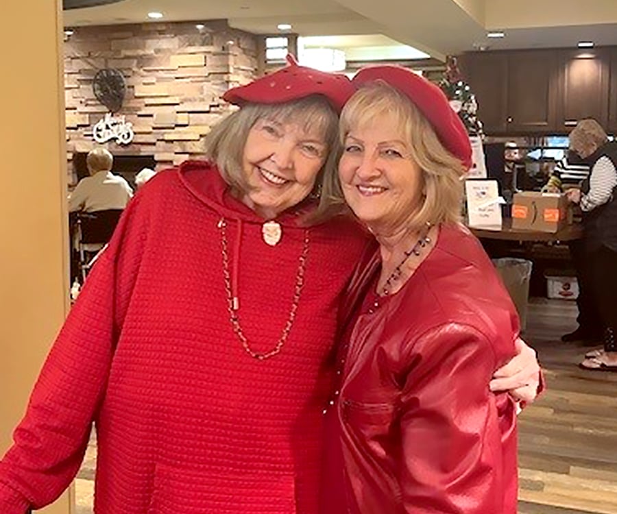 Two smiling older women wearing red outfits and matching red berets stand close together, embracing each other in a cozy indoor setting with people and festive decorations in the background.