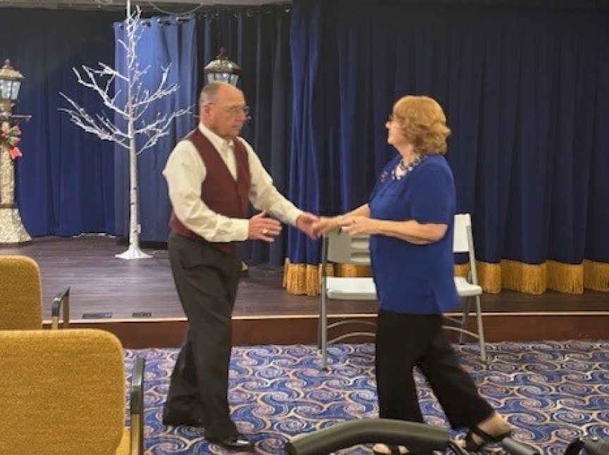 An older man in a maroon vest and an older woman in a blue top dance together in a carpeted room with blue curtains, gold-trimmed chairs, and a decorative bare tree on stage in the background.