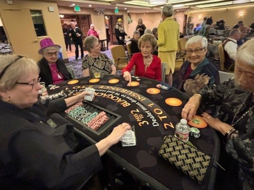 A group of elderly women sits around a blackjack table in a casino, some smiling and wearing festive attire. Chips and cards are spread on the table, and the atmosphere is lively and social.