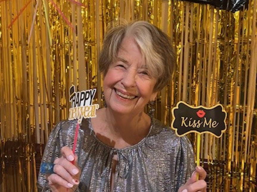 An older woman in a sparkly silver top smiles, holding signs that read Happy New Year and Kiss Me, standing in front of a shimmering gold tinsel backdrop.