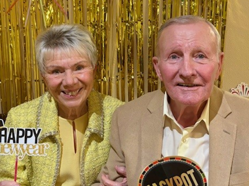 An older woman and man, both smiling, pose in front of gold tinsel decorations. The woman holds a Happy New Year sign, and the man holds a Jackpot sign. Both wear yellow-toned outfits.